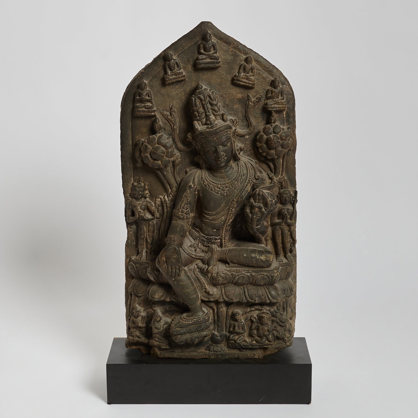 An Indian Carved Blackstone Stele of Avalokiteshvara Pala Period: Carved in deep relief to show the central figure of Avalokiteshvara seated at royal ease upon a lotus throne and displaying varada mudra; the peaked stele otherwise carved with smaller attending figur