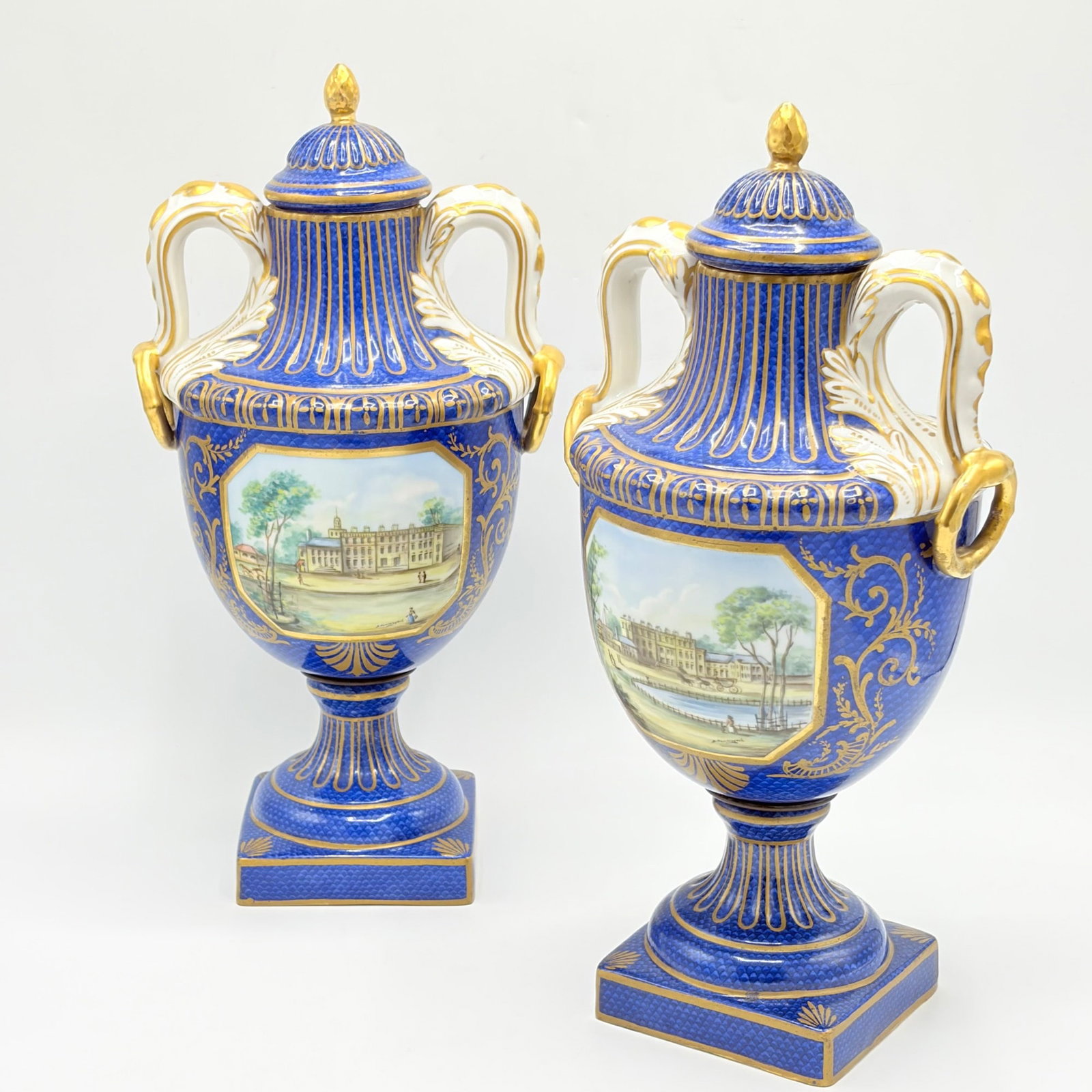 Pair of French Gilt and Hand-painted Porcelain Urns: Mid-19th century Each with domed cover, blue ground, fluted neck, loop handles and gilt faux rings, on a baluster form body with handpainted landscape reserve signed D. Pommeyrol, on a square base. Ea