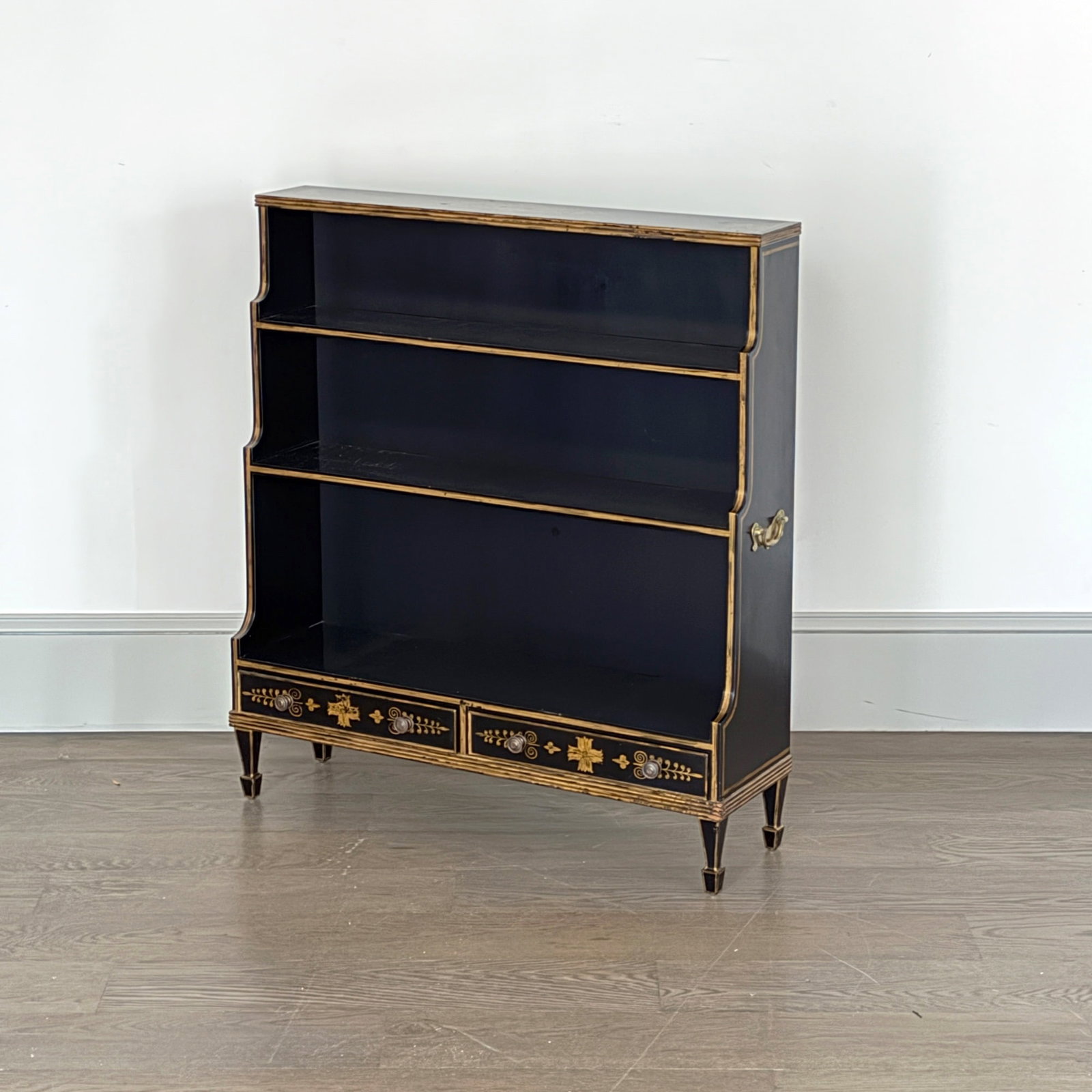 Regency Style Black Painted and Gilt Decorated Bookshelf: Mid 20th century The top with applied brass reeding, with three graduated shelves above two short drawers, all on square tapering legs ending in spade feet. Height 40 1/2 inches, width 36 1/2 inches, 