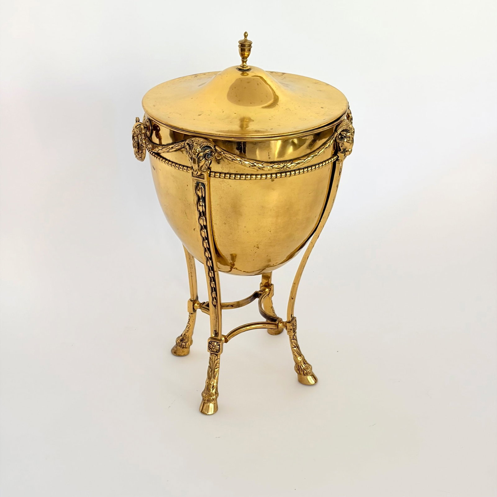 Neoclassical Brass Covered Urn: Late 19th century The lid with urn finial above the body with wreath ring handles and ram's heads around the rim, all raised on hoof feet. Height 25 3/4 inches, diameter 13 3/4 inches.