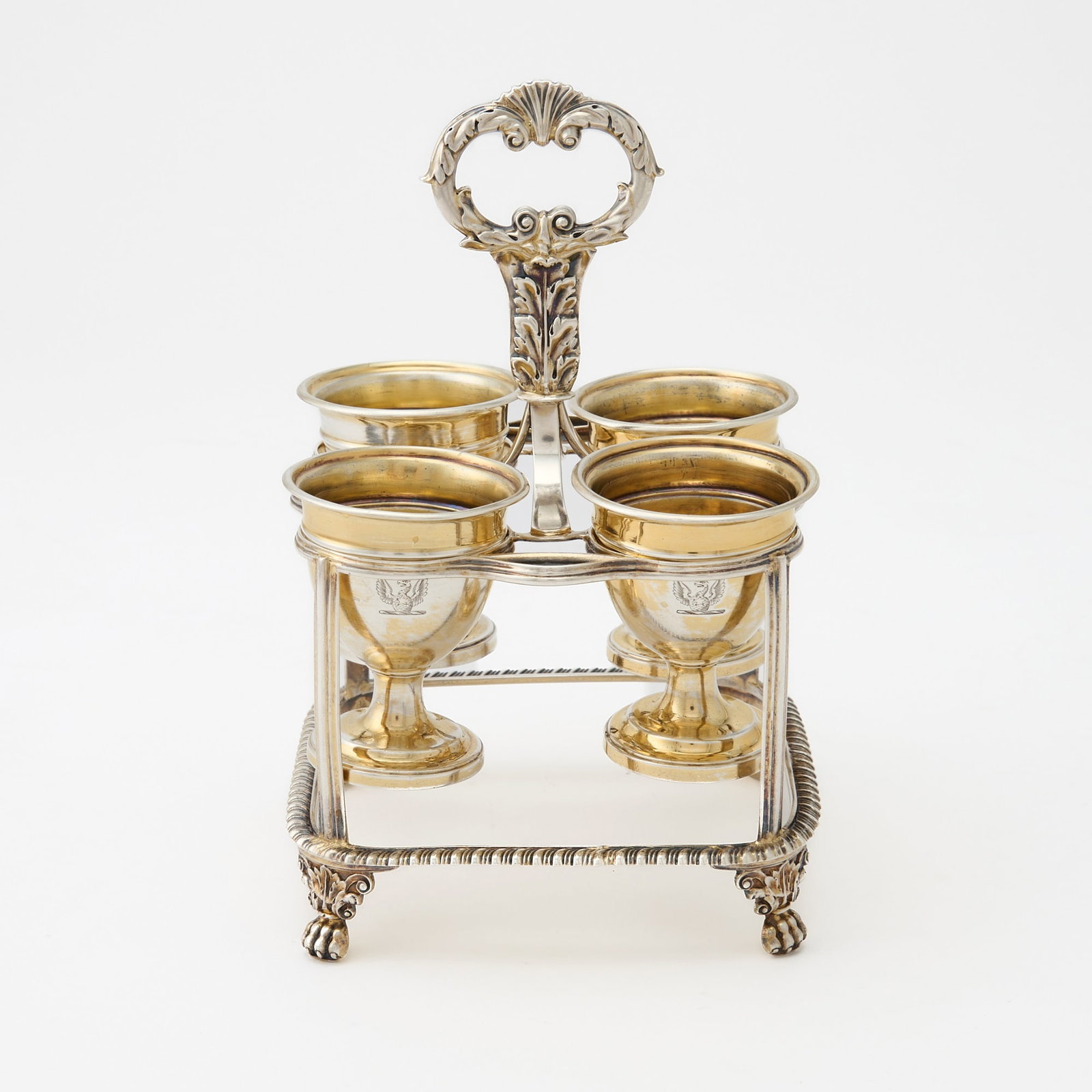 George III Sterling Silver Egg Frame: William Burwash, London, 1817 The square frame with gadrooned rim, fitted for four cups, with foliate ring handle, on four paw feet. Height overall 7 inches (18 cm), total approximately 13 ounces.