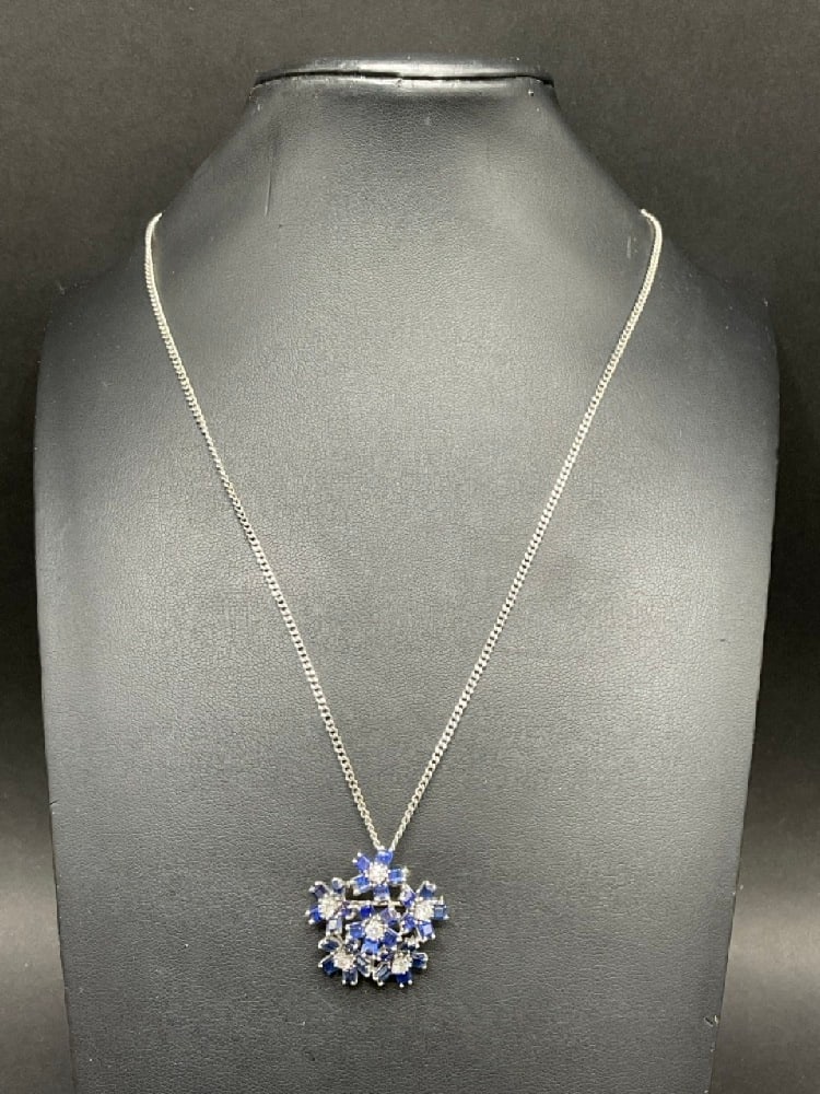 An 18K White Gold Diamond and Blue Sapphire Flower Pin/Pendant Necklace: An 18K White Gold Diamond and Blue Sapphire Flower Pin/Pendant Necklace. A floriform 1" pendant with six (6) diamonds and thirty (30) blue sapphire stones with 18K white gold chain. Total weight: