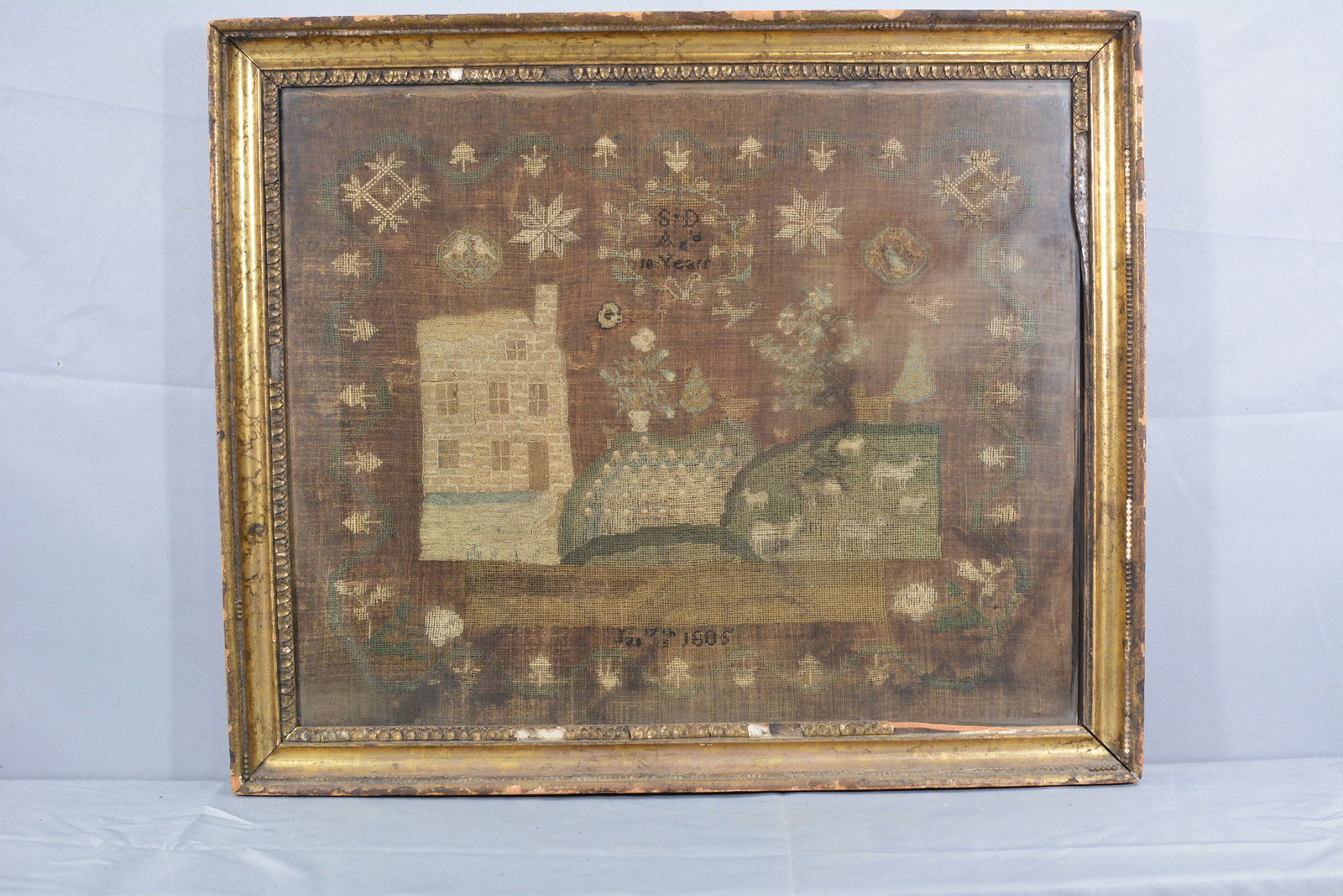 1805 Sampler: 1805 Sampler by S D 10 years. Needlework of a building, birds, sheep in a meadow in gilded frame. Some condition issues, needs a cleaning, 25"W x 21"T From the collection of Doug & Sandy Davies.