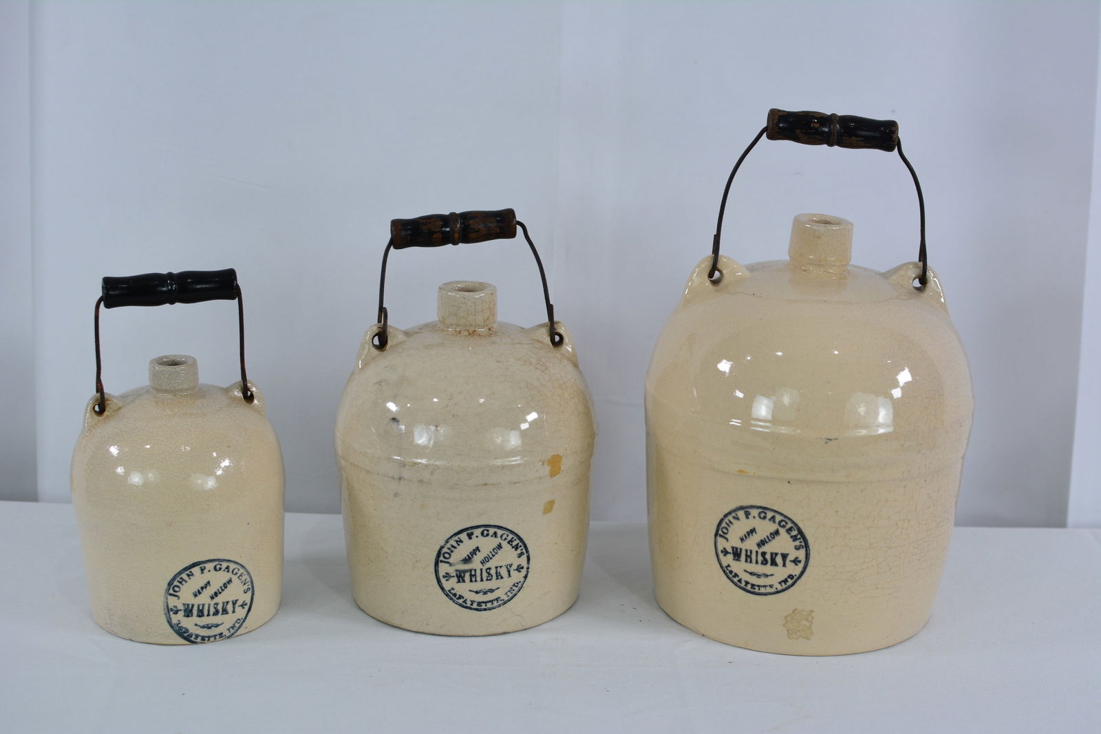 Three graduated size stoneware jugs w/ "John P. Gagen's Happy Hollow Whisky Lafayette, Indiana": Three graduated size stoneware jugs w/ "John P. Gagen's Happy Hollow Whisky Lafayette, Indiana" w/wire bail & wood handles.