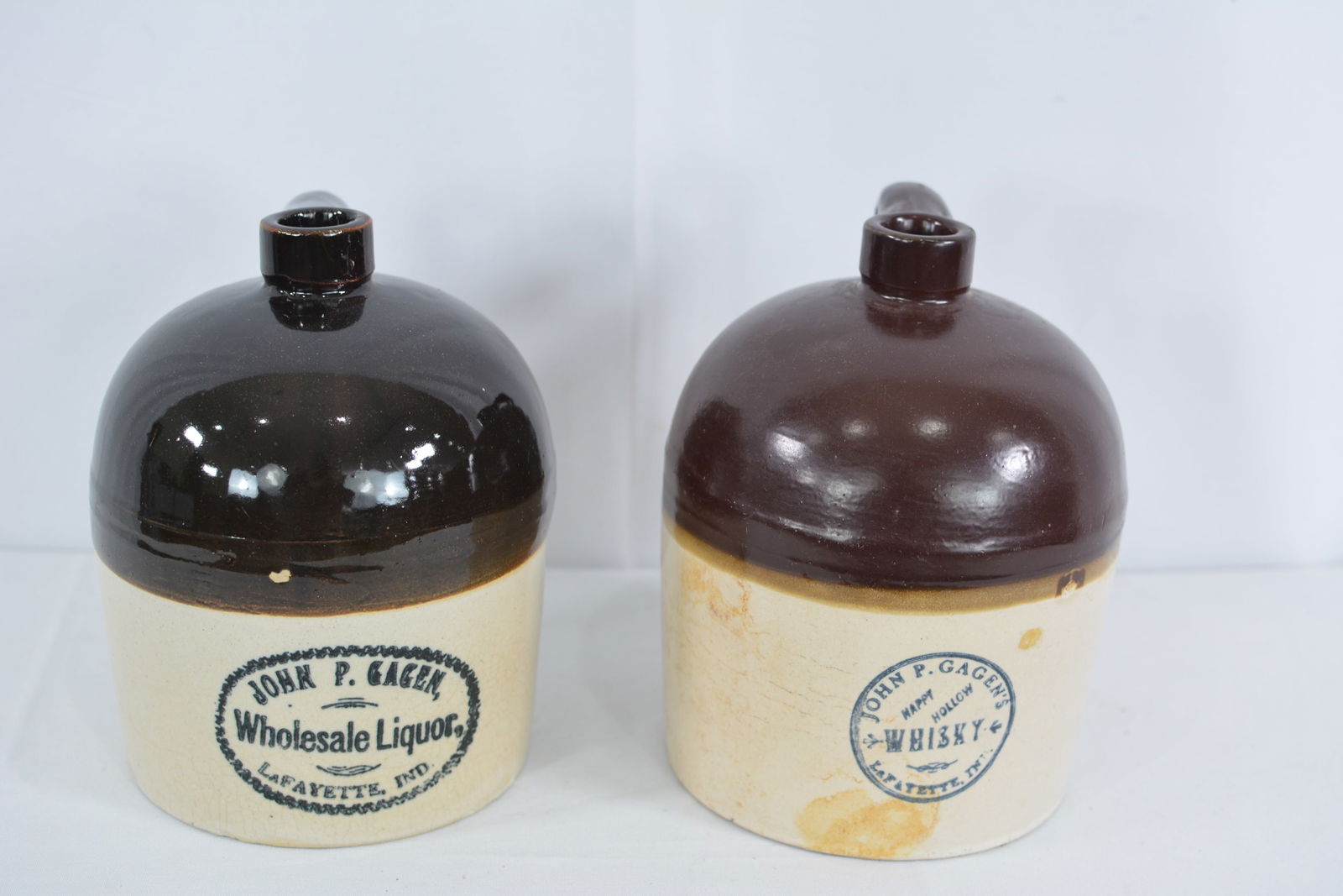 2 bicolor John P. Gagen stoneware jugs. One is Wholesale Liquor and one is Happy Hollow High Gap.: 2 bicolor John P. Gagen stoneware jugs. One is Wholesale Liquor and one is Happy Hollow High Gap. One has cracked handle. Both marked Lafayette Indiana.