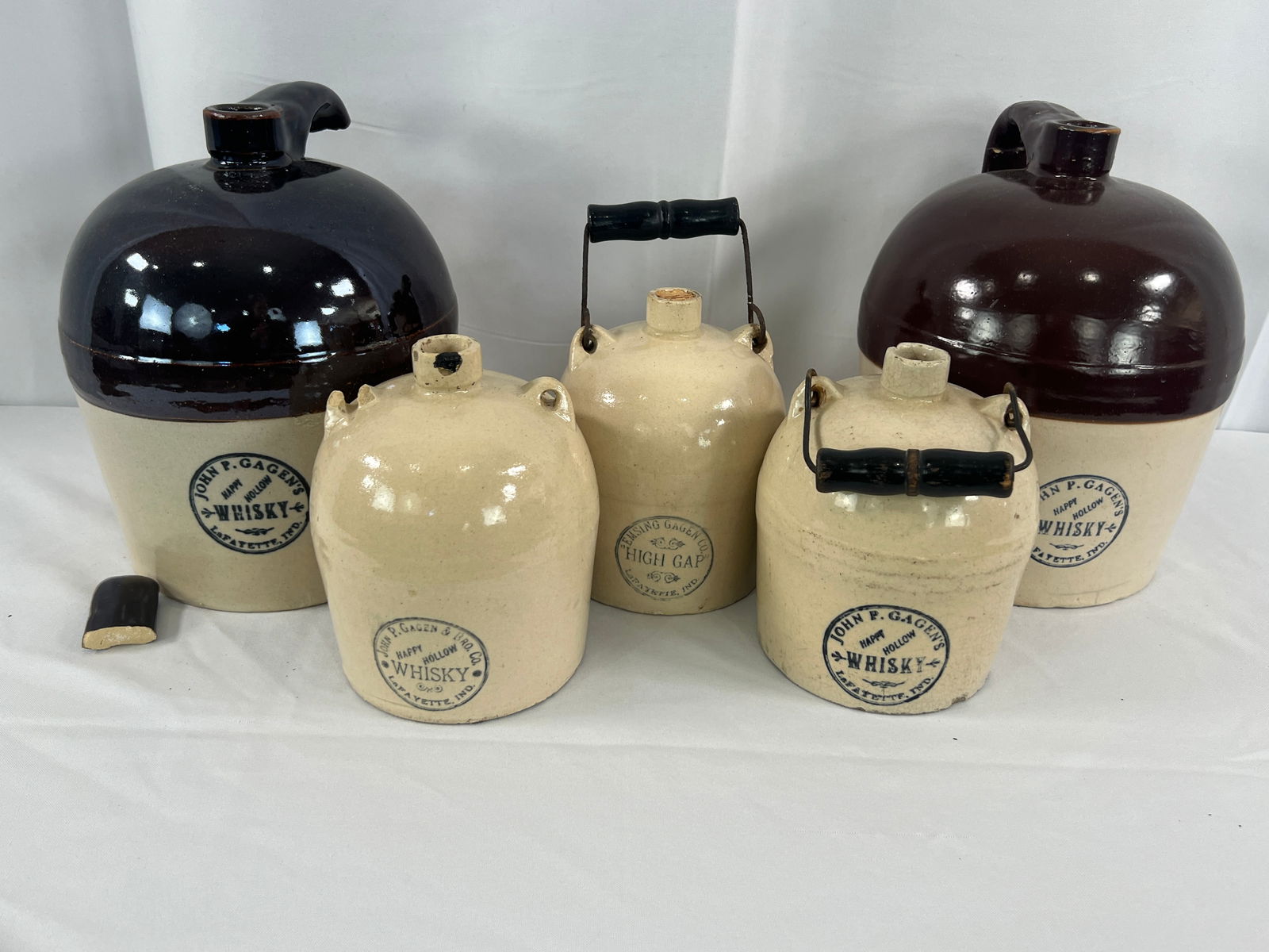5 Gagens Whisky Stoneware Jugs: 4 Gagens Whisky Stoneware Jugs, John and Emsing Hagen advertising whisky jugs, High Gap and Happy Hallow, Lafayette, IN, 2 bicolor gallon jugs, stoneware some with bail handles, hairlines and chips,