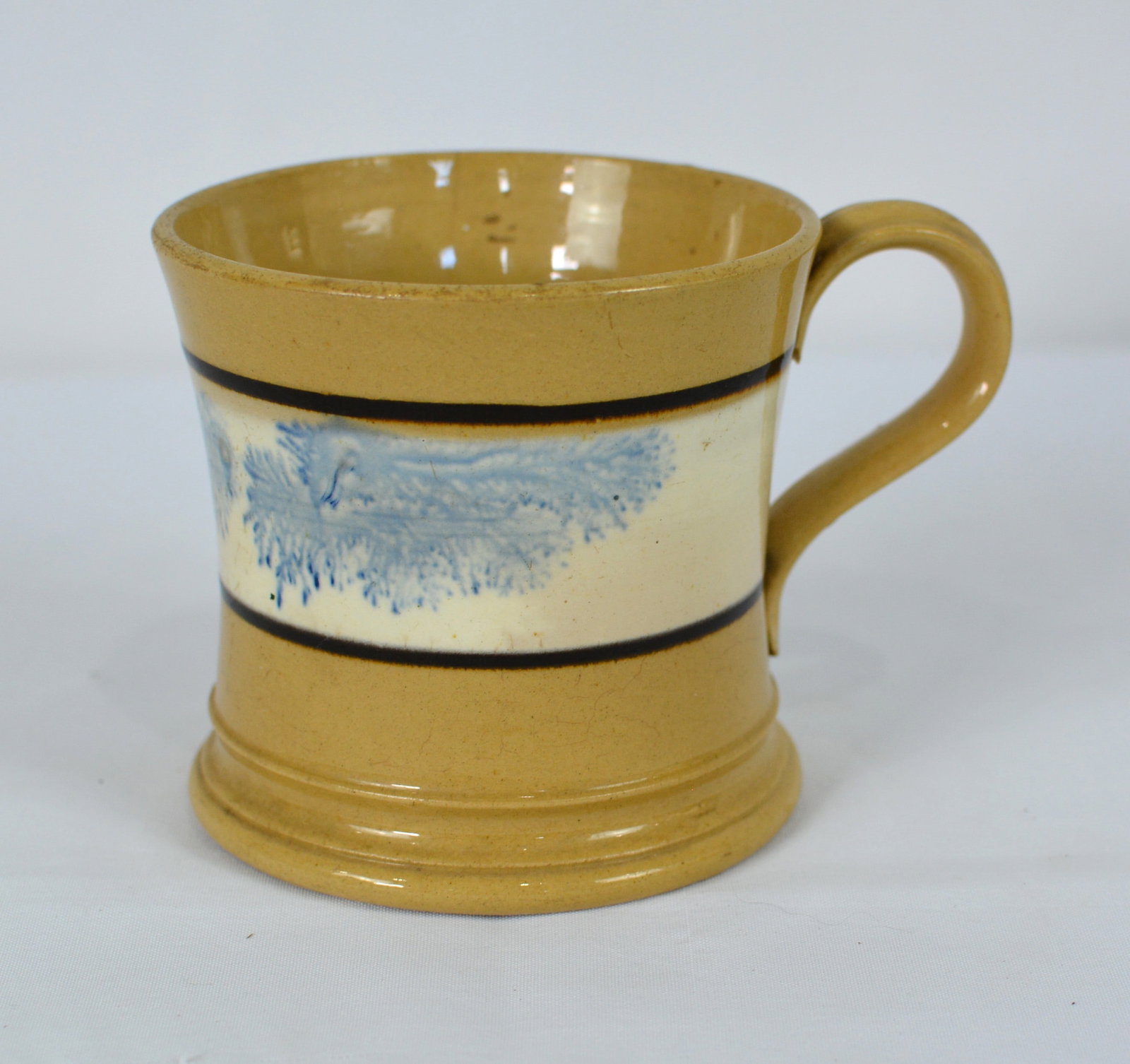 19th Century Mocha ware Tankard Mug with Blue Decoration: Early 19th-century English mocha ware tankard featuring a pale blue "seaweed" decoration centered within a cream-colored slip band and bordered by black bands. On a raised on a molded foot, with an