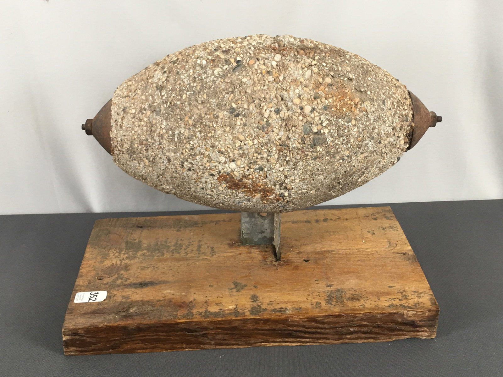 Concrete Windmill Weight: Concrete Windmill Weight, football shaped, on wooden stand. 18"L x 9.75"W x 13"H. **Please note this lot will be very heavy to ship.**