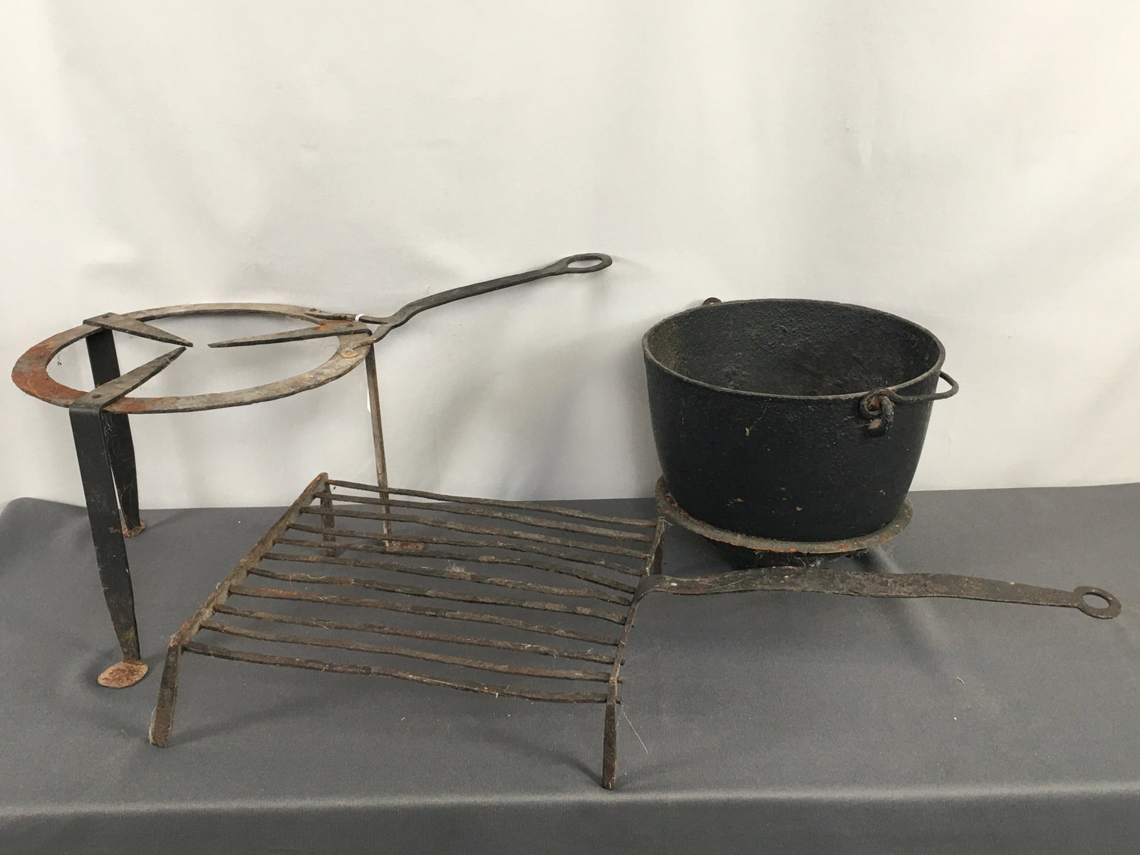 Fire Trivets, Cast Iron Pot: (2) Wrought iron fire trivets, square and round, with long handles. 3-legged cast iron kettle. Square trivet 25.5"L w/ handle x 11.5"W. Round trivet 20.5"L w/ handle x 11"W. Kettle 10"D x 7.75"H.