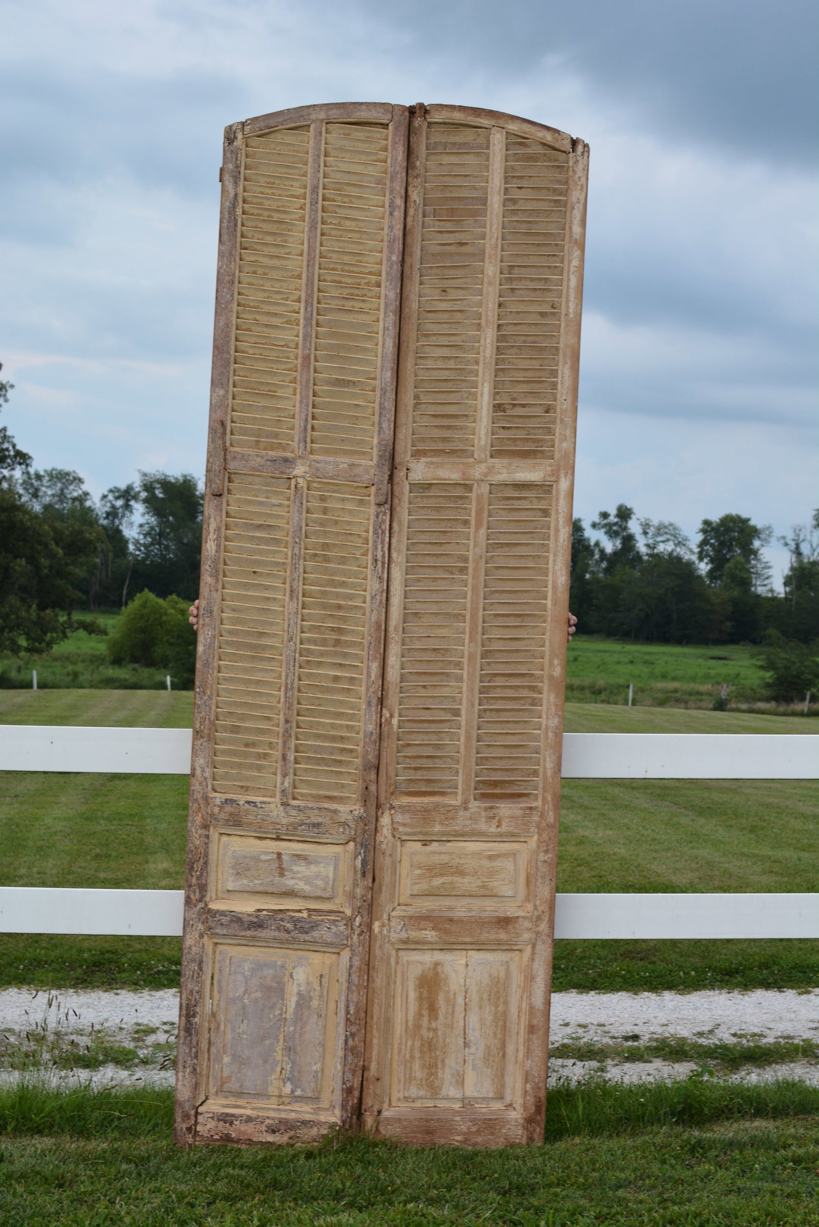 Pair of Tall 19th Century Architectural Louvered Shutter Doors: Impressive pair of tall 19th century architectural wooden shutter doors featuring arched tops, extensive louvered sections, and raised lower panels. Retain layers of old weathered buttermilk paint