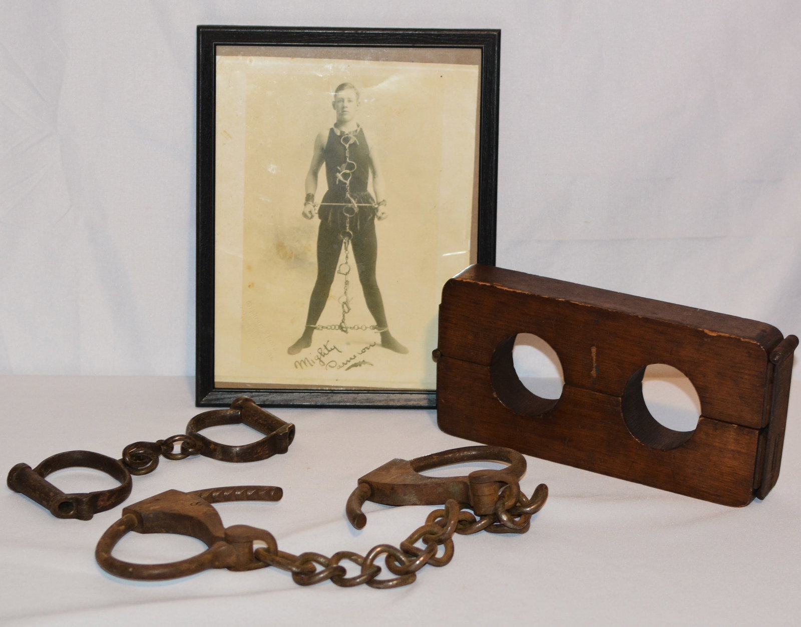 Antique Magician / Escape Artist Restraints with Mighty Samson Photograph: Unusual collection of antique escape-artist or magicians restraints consisting of two sets of iron shackles and a heavy wooden double-hole restraint or stock with hand-forged metal hinges and locking