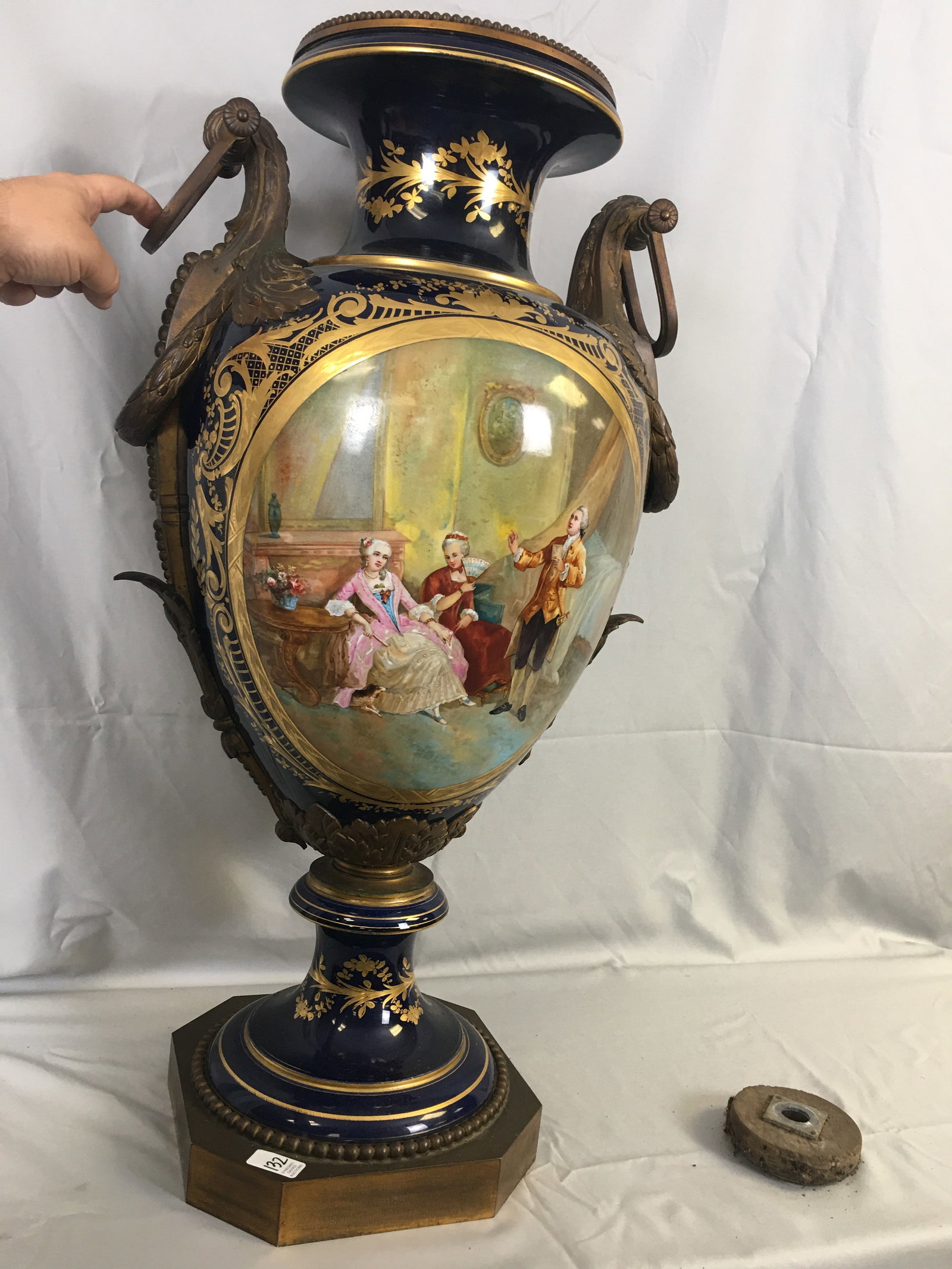 Hand Painted Urn on Pedestal: Hand Painted Porcelain Urn on Pedestal. In navy blue with gilt detailing, having brass handles & leafy accents. Front side features Continental gentleman & ladies, reverse shows trees in a lake.