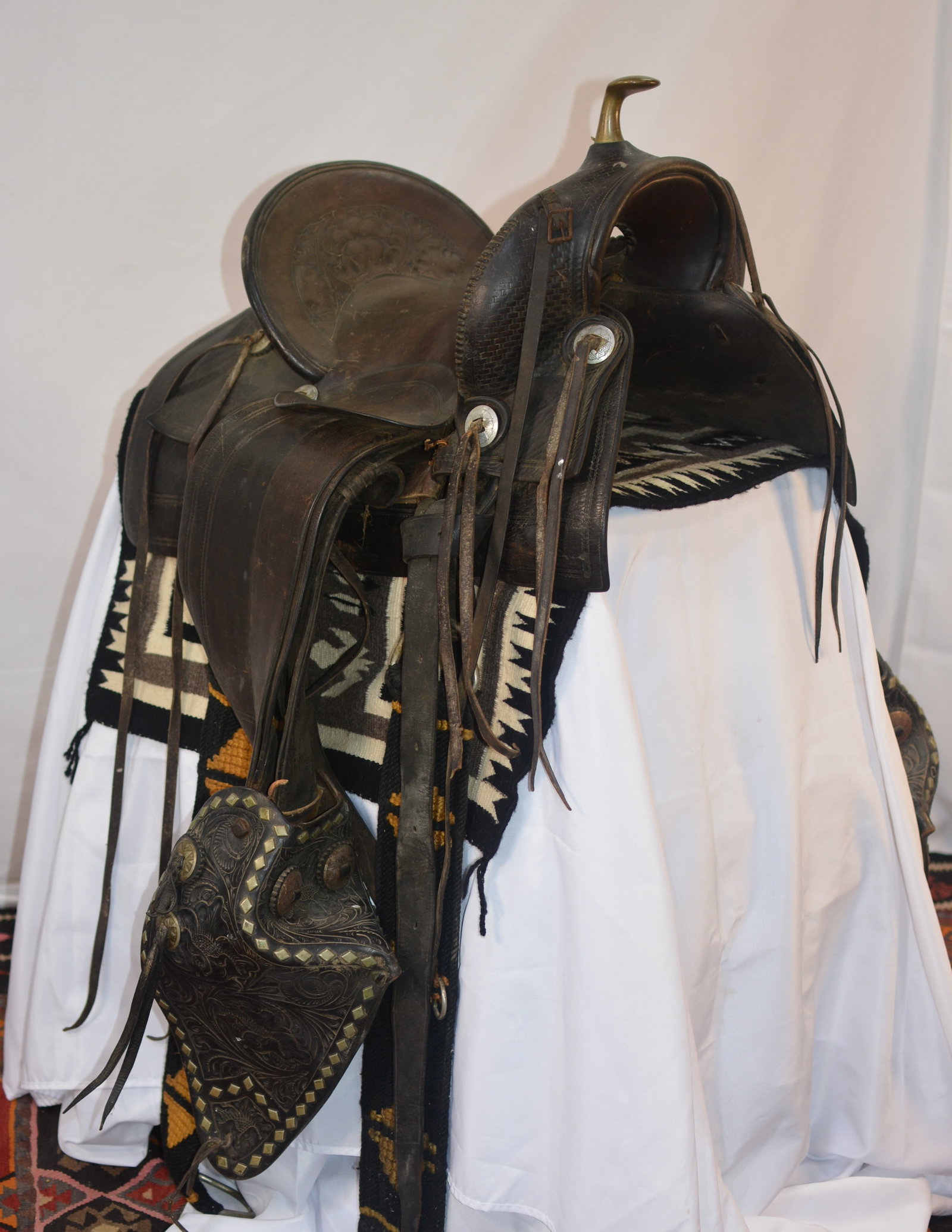 Antique Western High-Back Saddle with Tooled Tapaderos: American Western high-back saddle featuring a prominent brass horn, high cantle, dark leather construction, and decorative basket-weave tooling. Accompanied by an impressive pair of large tooled