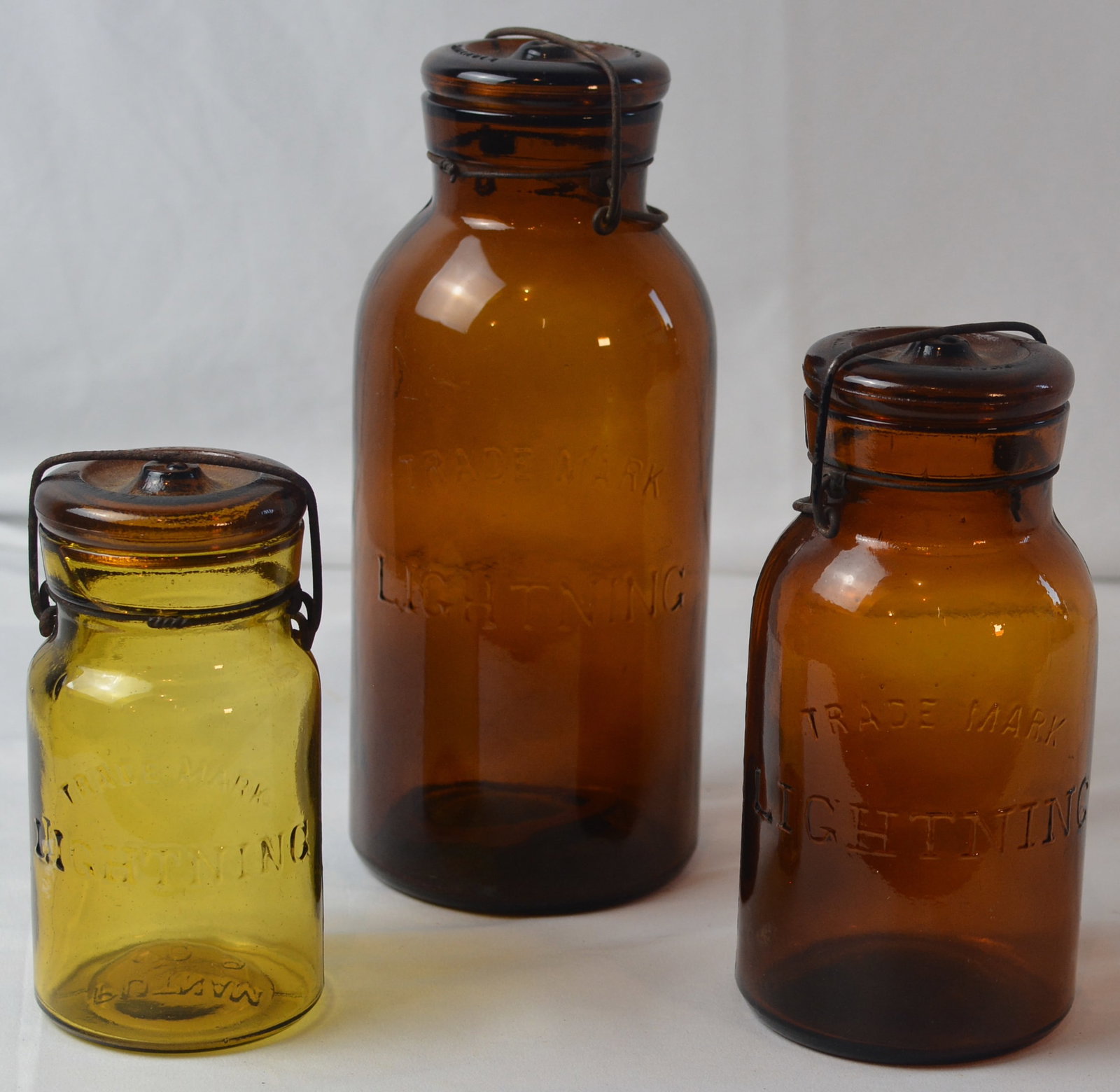 Three Lightning Putnam Trade Mark Glass Canning Jars with Bail Lids: Group of three early glass canning/preserve jars embossed TRADEMARK LIGHTNING, including two amber examples and one less-common Honey Amber example. Each features the characteristic glass lid with wir