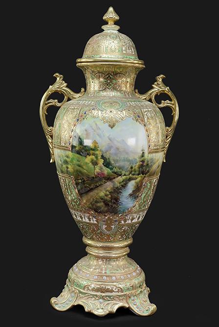 A Nippon Porcelain Covered Urn.: A Nippon Porcelain Covered Urn. Double handled porcelain urn with a painted scene of mountains and water on either side. Urn is further decorated with gilding and enameling throughout Height: 38"