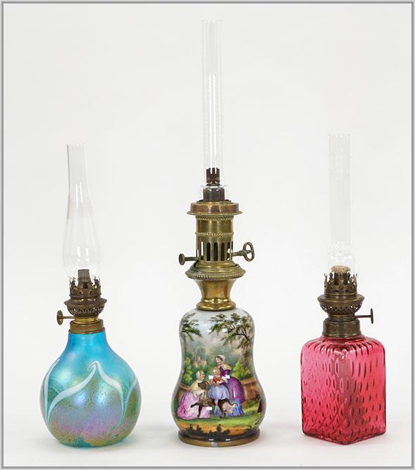 Three Contemporary and Antique Oil Lamps.: Three Contemporary and Antique Oil Lamps. Consisting of a cranberry diamond quilted Victorian oil lamp. Together with a colonial scene porcelain oil lamp and a contemporary Steven Bradshaw 1984 irides