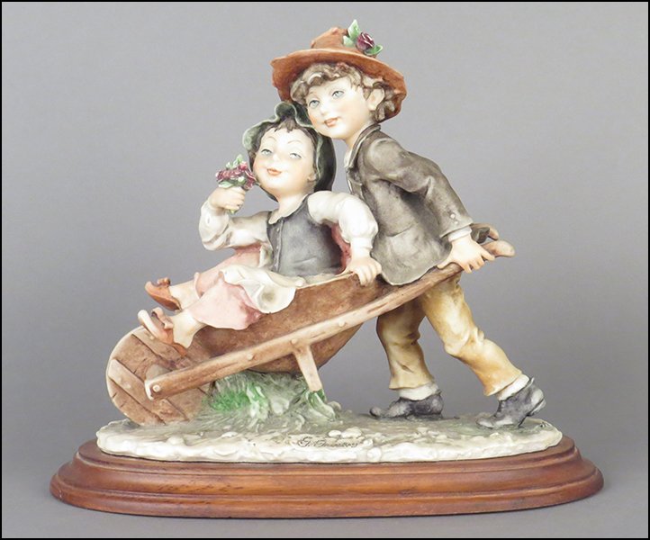 GIUSEPPE ARMANI 'BOY PUSHING WHEELBARROW WITH GIRL': GIUSEPPE ARMANI 'BOY PUSHING WHEELBARROW WITH GIRL' FIGURAL GROUP. Signed on the sculpture. Copyright Florence 1982. Bearing the Capodimonte 'N' crown stamp. Raised on a wood base (1'' x 11'' x 5.25''