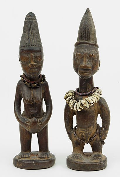 A Pair Of African Carved Wood Ibeji Figures.: A Pair Of African Carved Wood Ibeji Figures. One male, one female. Male figure bears cowrie shell necklace. Female figure bears metal chain necklace Height: 12"