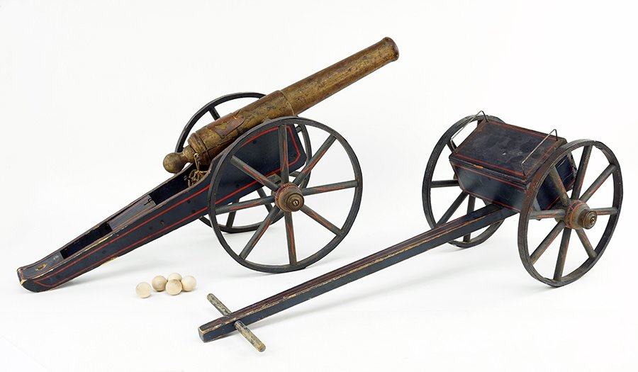 A Toy Cannon.: A Toy Cannon. Late 19th / Early 20th Century painted wood toy cannon with metal rimmed wheels. Cannon is decorated with a tooled leather eagle. Cannon is accompanied by a pull cart with attached box c