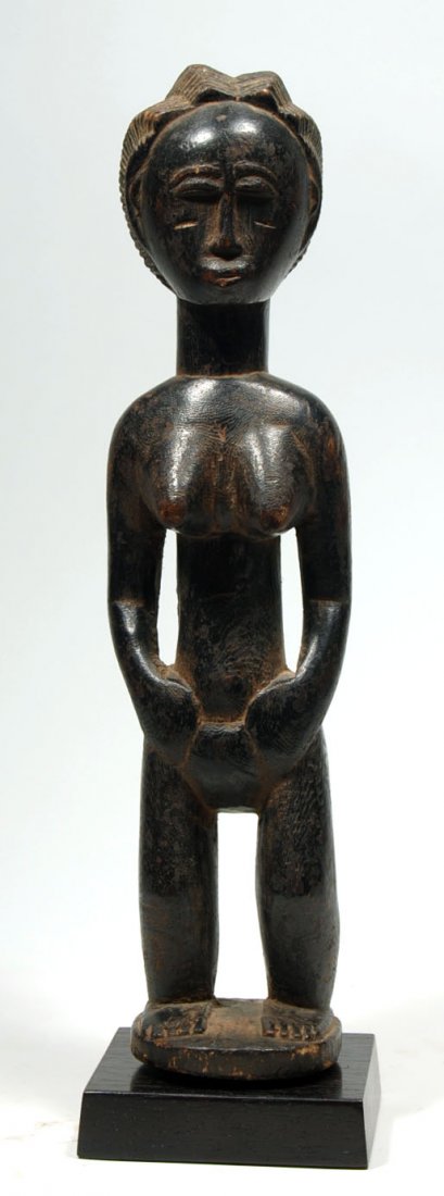 African Baule Figure: Baule figure from Ivory Coast, wood, standing on integral base, hands on hips, with small jutting breasts, tubular neck, and tribe typical facial features including arched brows, thin nose, semi-circu