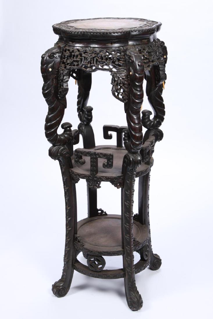 HEAVILY CARVED TEAK AND LOBED (2) SHELF PEDESTAL: Carved and pierced prunus flowers throughout. Figural elephant heads with trunks double as legs and support the top from the middle shelf with T -form rails. Lower shelf is directly above circular str