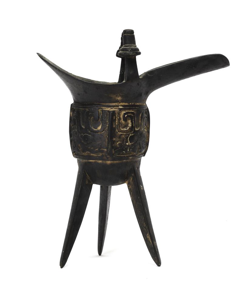 ARCHAIC CHINESE SHANG DYNASTY BRONZE JUE: An ancient Chinese vessel used to serve warm wine during ancestor-worship ceremonies. Cast with spout, (2) figural handles and band of raised geometric patterns. 7 inches