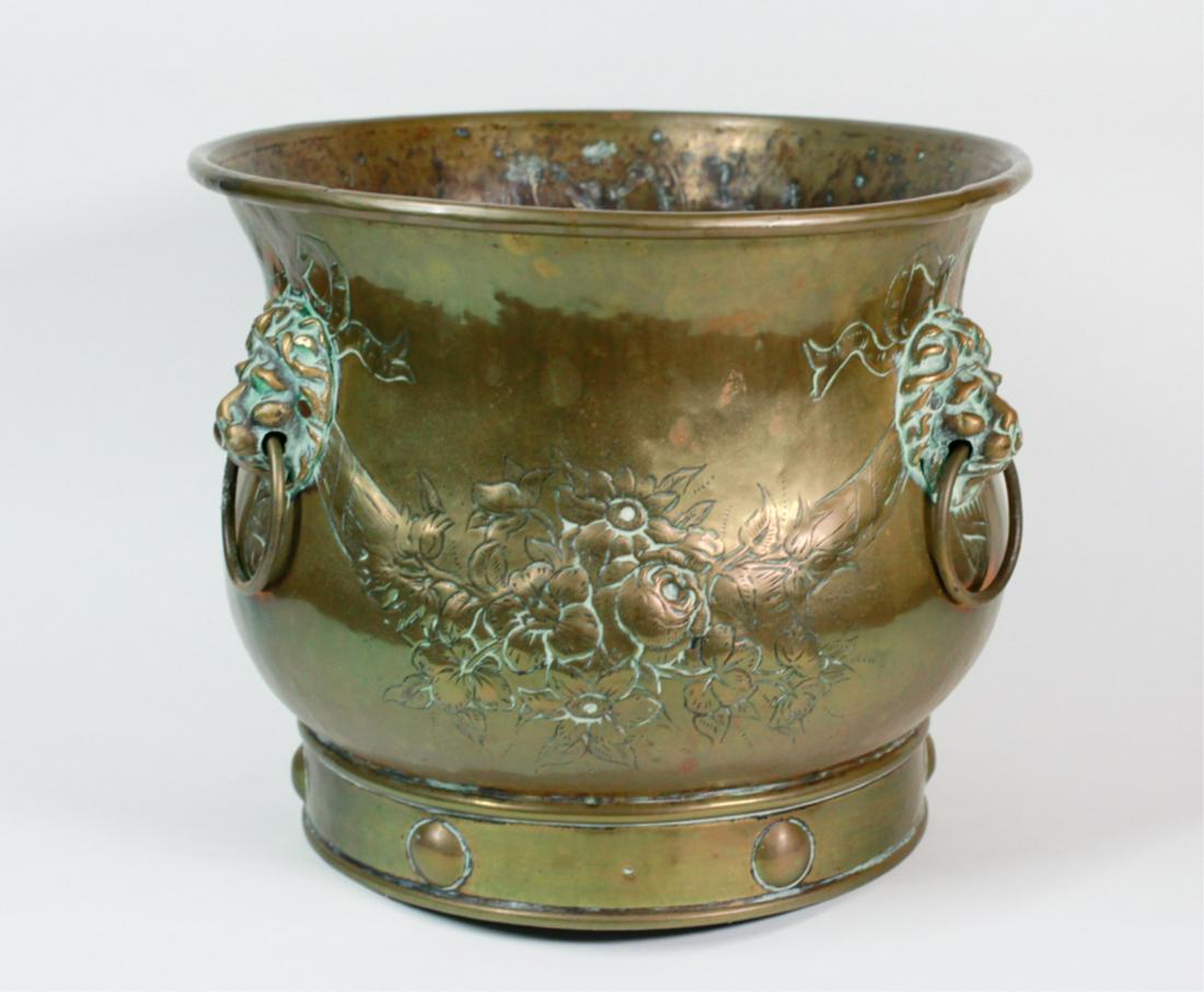 GEORGE III HAMMERED BRASS JARDINIÈRE: Flared rim, mounted with three ringed lion masks, each joined by engraved and chased ribbon and bow swags centered by flowers and one with a pineapple and fruits, on a ringed base with circular relief