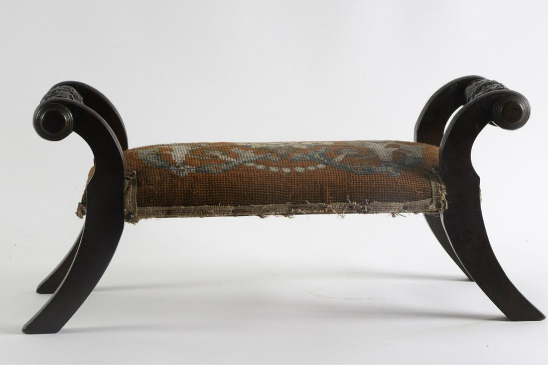 FINELY CARVED MAHOGANY UPHOLSTERED FOOTSTOOL: Sabre legs with scrolled arms carved with thistles and volute returns. Upholstered in needlepoint with a floral motif. 9 1/2 x 20 x 8 1/2 inches. . Condition: Repair to breaks on (1) arm. Lamination e