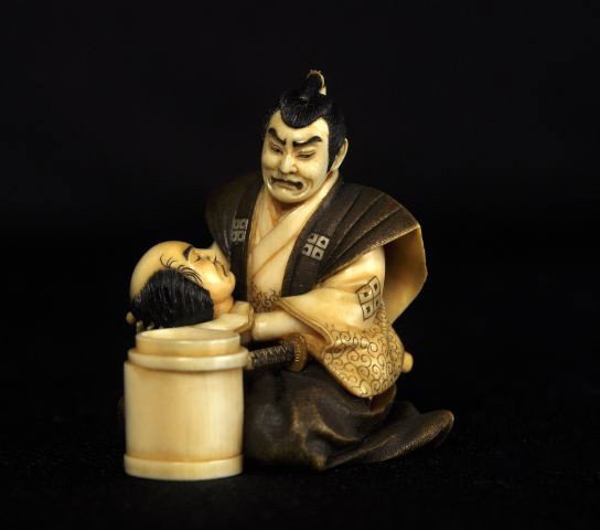 Japanese 20thC Tinted ivory figural netsuke: Japanese 20thC Tinted ivory figural netsuke, carved as a samurai warrior in formal dress and about to place a severed head of an enemy in a container, signed Masatoshi? (possibly? Nakamura Tokisada, 1
