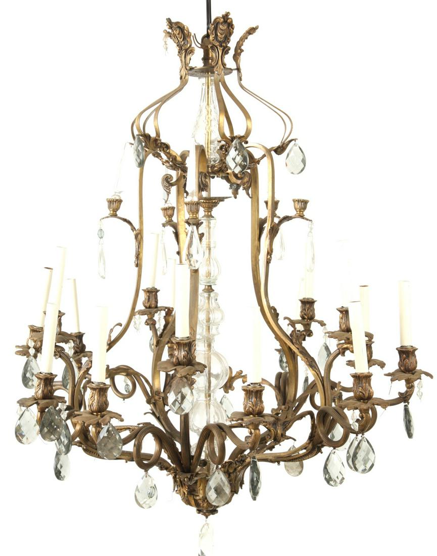 ROCOCO STYLE GILT BRASS (15) LIGHT CHANDELIER: (15) semi-posable arms surround a central brass column that passes through a decorative glass shaft. Adorned with acanthus leaf motif, smoke glass teardrop prisms, prism obelisks, and a central double