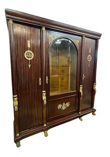 GILDED AGE ERA MAHOGANY BOOKCASE w/ ORMOLU MOUNTS: Molded entablature surmounts a (3) door case with a center-arched beveled glass door that is flanked by (2) ormolu mounted examples. Each cabinet door is separated by (4) demilune columns with figural