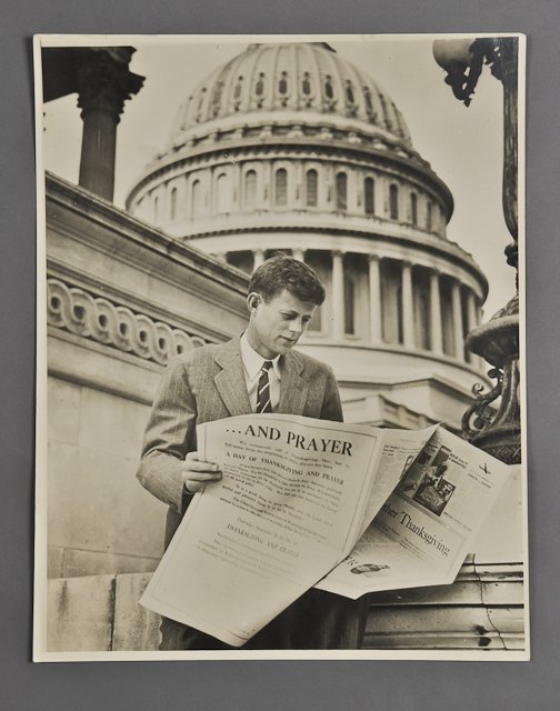 1946-John F. Kennedy, Photographic Portrait: (1) Photograph used for promotional material of a young J.F.K. reading a news extra regarding Thanksgiving and prayer in front of the United States Capitol Building. 14" x 11 1/2". Condition: Toning