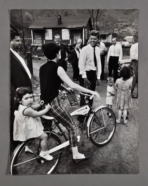 1960-May, J.F.K. Campaigning in West Virginia: (1) Large format photograph for "Life" Magazine of J.F.K. speaking with a woman on bicycle, 10 1/2" x 13", taken by Hank Walker. Condition: Crease, lower right