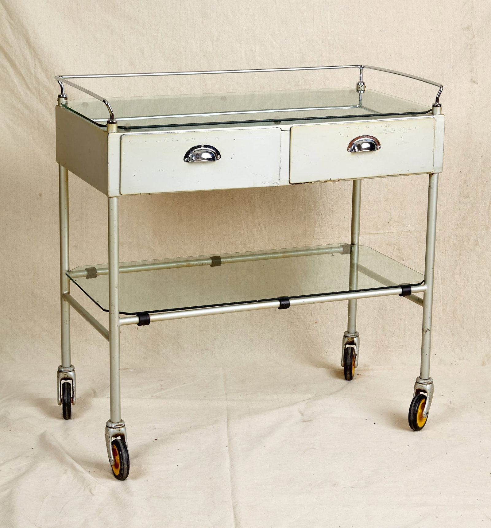 MID-CENTURY HOSPITAL TROLLEY: Unmarked. Chrome gallery surrounds glass top, Structural steel (2) drawer case with lower glass shelf is raised on casters. 37 x 36 1/2 x 18 1/2 inches