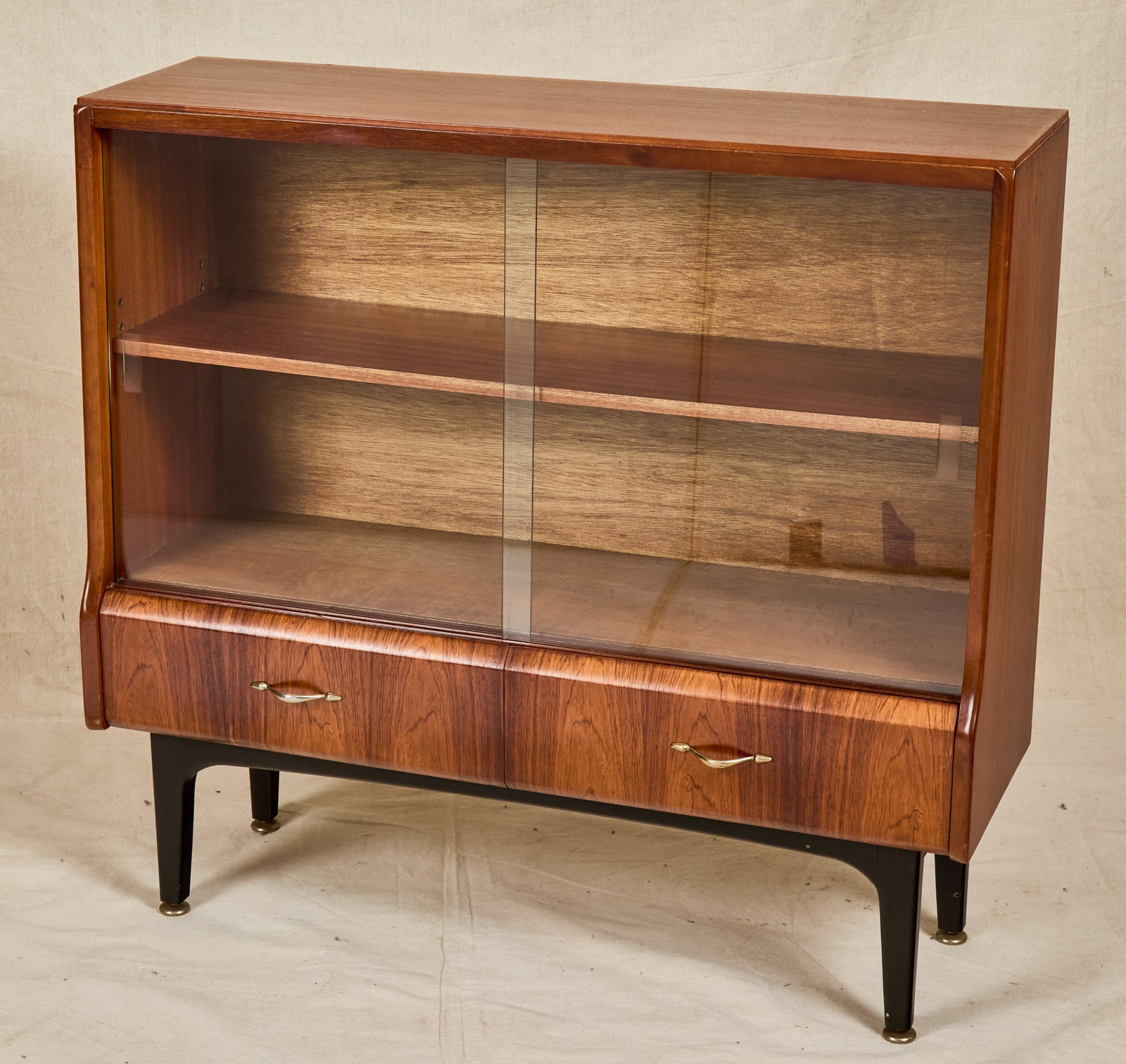 ROTHWELLS MID-CENTURY SLIDING GLASS DOOR BOOKCASE: Upper section with sliding glass doors and single shelf over (2) molded drawers with beautiful veneer, raised on a black painted base. 34 x 36 x 12 inches.