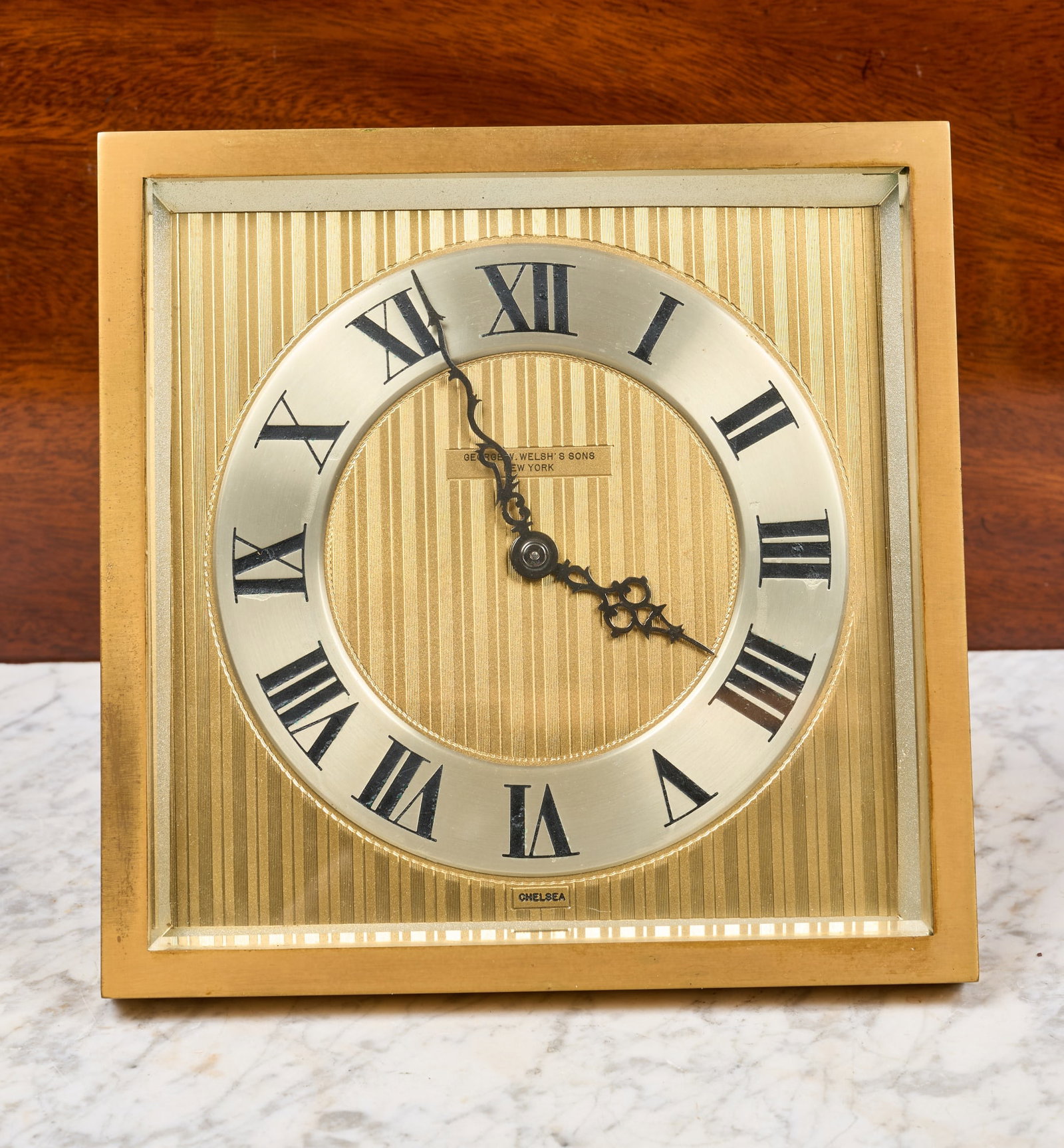 GEORGE W WELSHS & SONS CHELSEA DESK CLOCK: 8 inch brass square case with beveled glass, 5 1/2 inch silver dial with fancy hands on engraved brass background with George W. Welsh's & Sons (New York) retailer. Serial no. 184863. 7 x 8 x 4 1/4 in