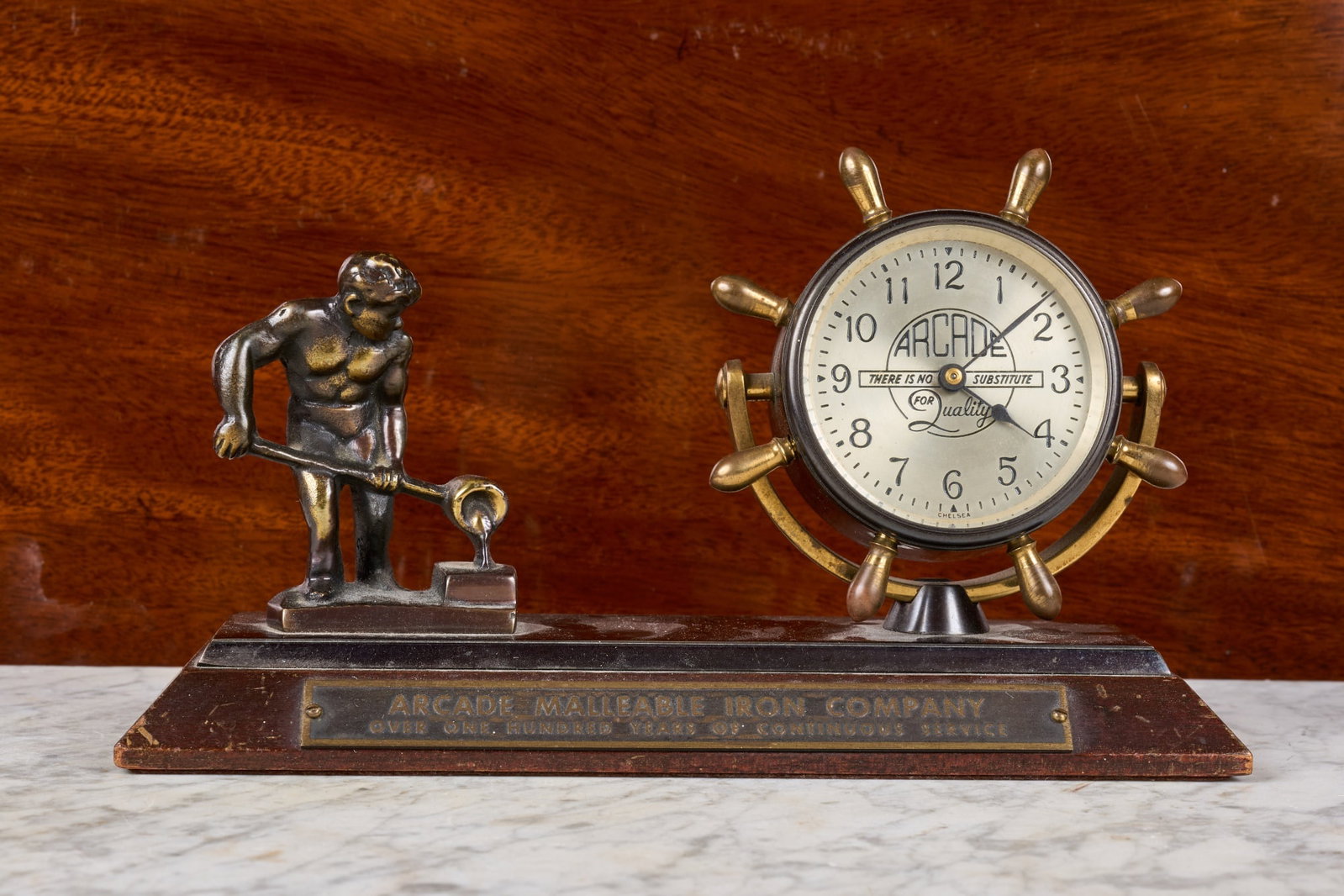 CHELSEA CLOCK CO. CUSTOM PRESENTATION DESK CLOCK: Chelsea Clock Co. bronze ships wheel clock with 2 1/4 inch silver dial engraved with Arcade tagline, on wooden base with bronze figure of an iron worker with crucible, and brass presentation plaque "A