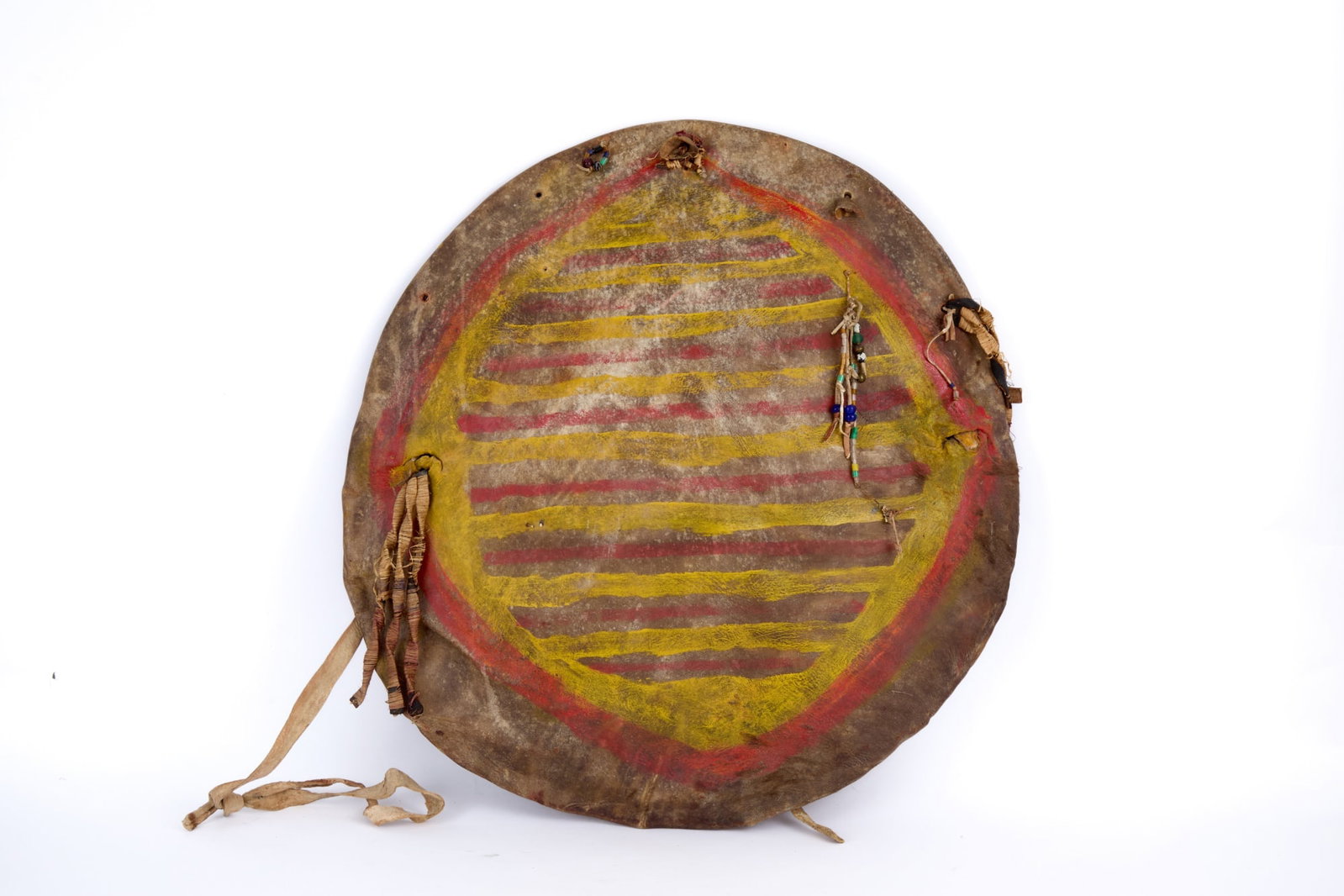 PLAINS INDIANS PAINTED RAWHIDE GHOST DANCE SHIELD: Plains Indians rawhide shield with polychrome painted sun/turtle shell?, bison dewclaw & bead rattles, grass wrapped leather tassels. 19 1/2 inches.