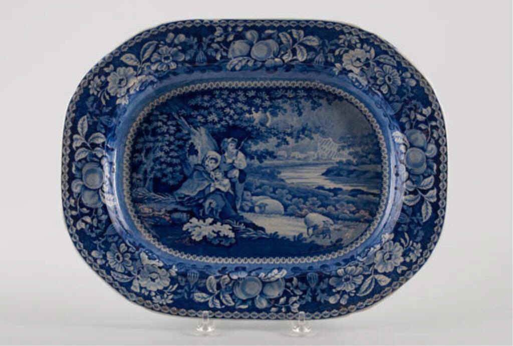 17-Inch Staffordshire Platter: Unsigned blue and white platter depicting a pastoral scene of a young couple and child under a large tree, sheep, water, church in background. 13 1/2" x 17". Condition: Good.