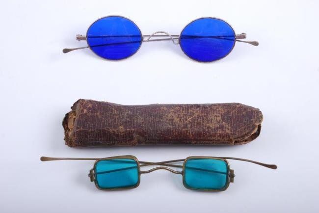 (2) 19thc. Spectacles with Tinted Lenses: (1) Rectangular brass frame with green tinted lenses, clear side lenses and paperboard case, (1) Oval steel framed example with blue tinted lenses. Condition: Corrosion to steel frames