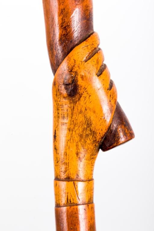 CARVED WOOD CANE WITH HAND HOLDING STAFF: Woodmont Conn"" knob handled cane carved from a single piece of wood includes a carved cuffed hand holding a ""stick"" with a carved alligator. Length 34 inches