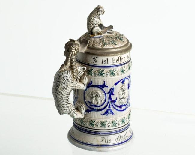 STONEWARE STEIN WITH CATS, MONKEY AND BEAR HANDLE: Forest green and cobalt highlight scrolls and leaves which surround animated cats. Bear handle supports pewter lid with stoneware monkey holding boot. Height 9 inches