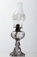 LIGHT AMETHYST PRESSED GLASS KEROSENE LAMP: Font is embossed with ring of bulls eyes. Lobed square base is embossed with ellipses, beads and acanthus leaves. Chimney is decorated with a band of frosted torches and laurel wreaths. Height 10 inch
