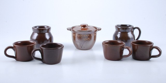 (7) Pieces of Brown Glazed Dorchester Pottery: Including: (1) 3 1/4 inch "Doe, Sullivan & Co. 1829" butter crock with lid, (1) 3 inch handled jug (lid absent), (1) 3 inch wide mouthed jar and (4) 2 inch cups. Condition: Good, handled jug missing l