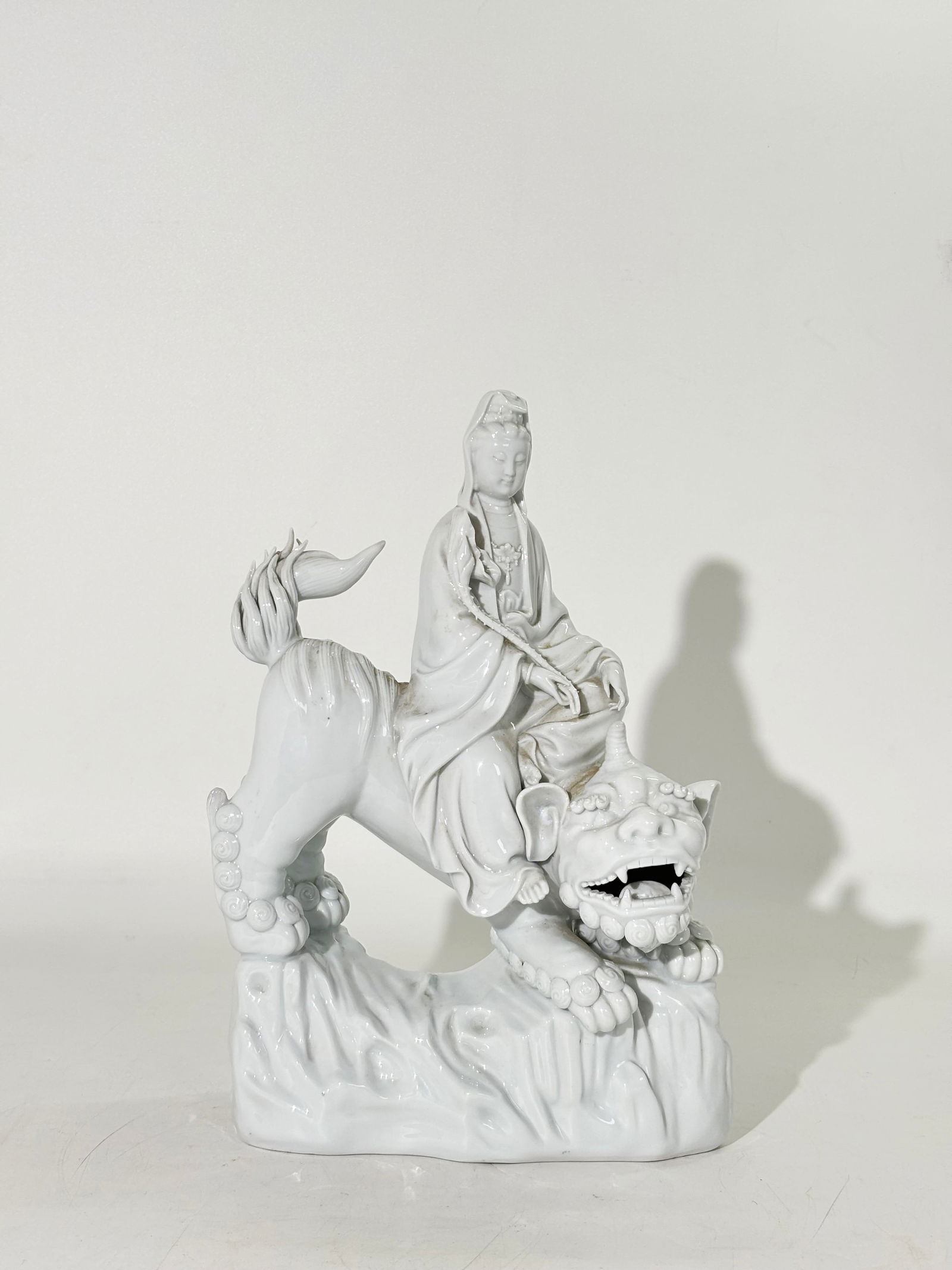 Chinese Blanc De Chine Guanyin Riding a Bixie: Chinese Blanc De Chine Guanyin holding a Lotus Bud & Leaf and Riding a Bixie with a rock-work base; the concave bottom glazed with wide unglazed foot. Height 11 1/2 inches X Width 7 3/4 inches.