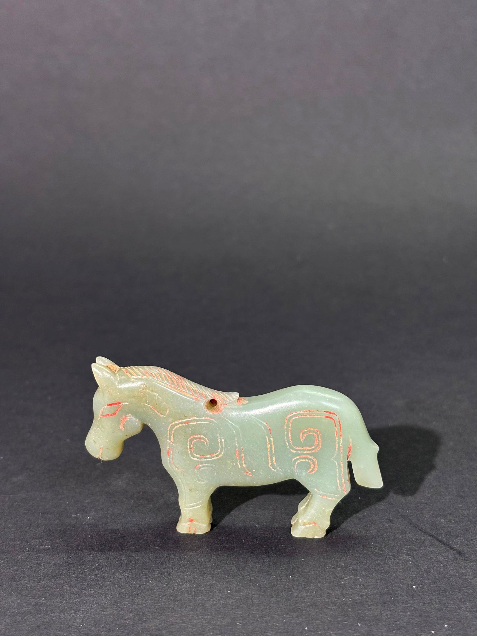 Chinese Celadon Jade Archaistic Horse Pendant: Chinese Celadon Jade Archaistic Horse Pendant with the body detailed in Shang Type lines now filled with slight red coloring and dirt; an irregular hole to base of mane for suspension. Height 1 5/8 in