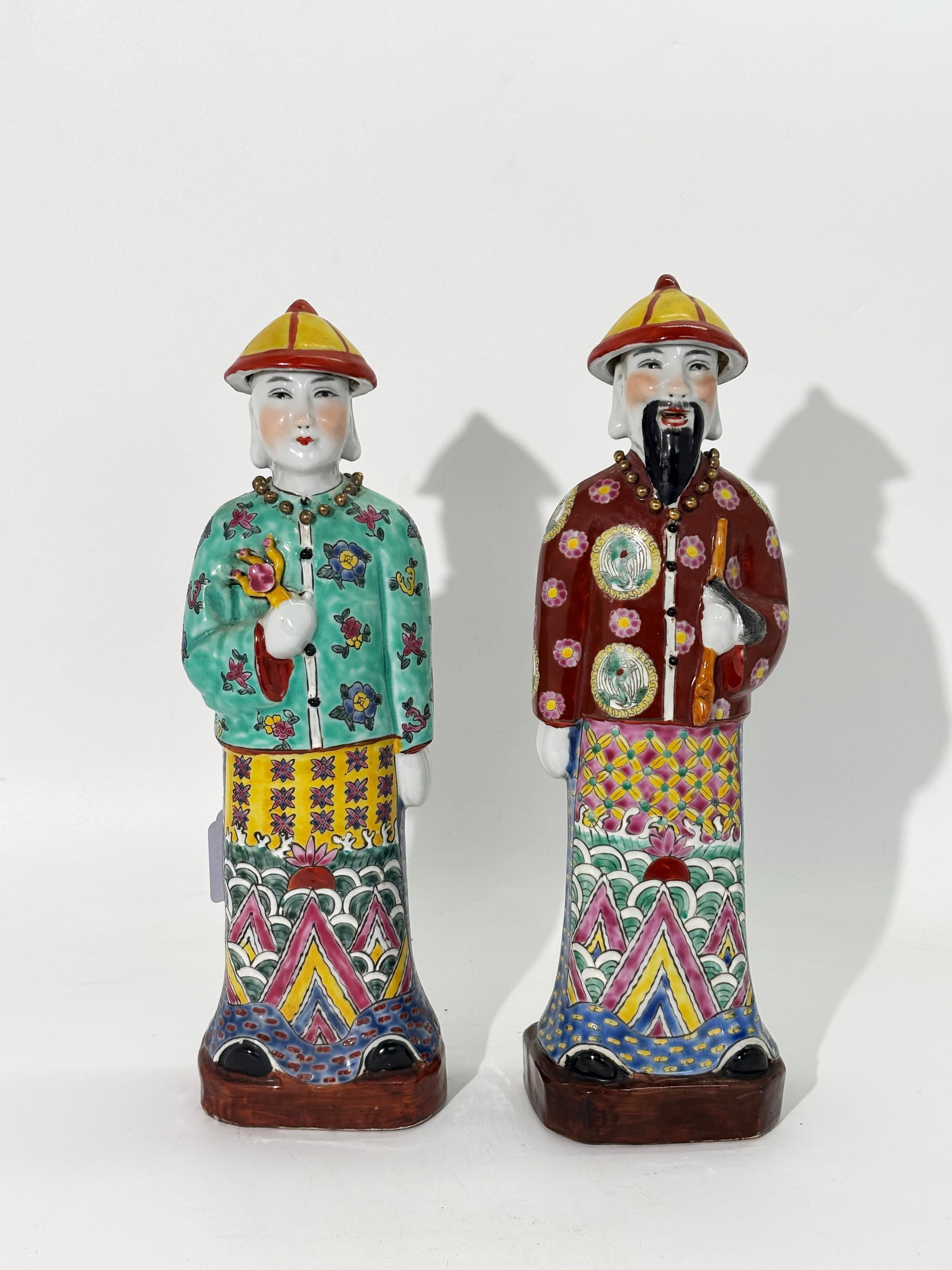 2 Chinese Porcelain Standing Manchu Officials: 2 complimentary Chinese Porcelain Manchu Officials with their hair in Manchu Queue pigtail and wearing Hat, gilt beads and elaborately patterned gowns each on matching brown glazed integral stands wit