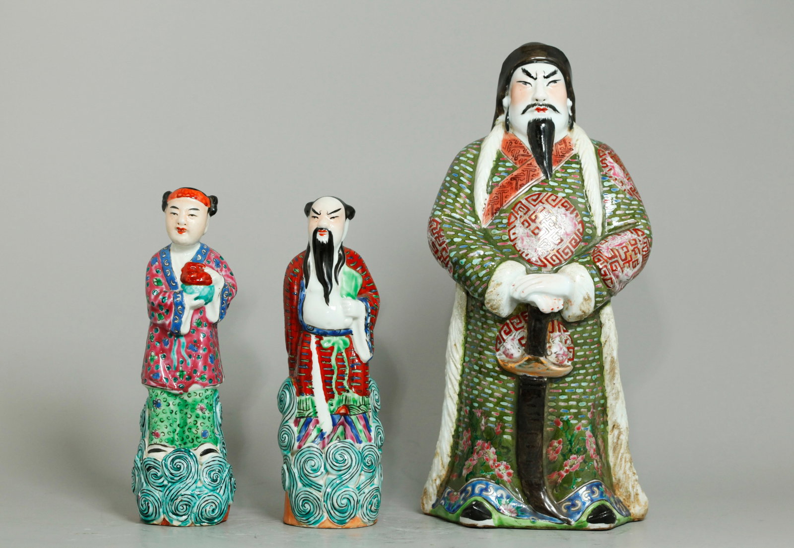 3 Chinese Enameled Porcelain Guardian Figures: 3 Chinese Enameled Porcelain Figures: the largest possibly Guan Yu with a white face and fur lined coat under a travelling hat over matching elaborate robes and with a large sword, the base unglazed; 