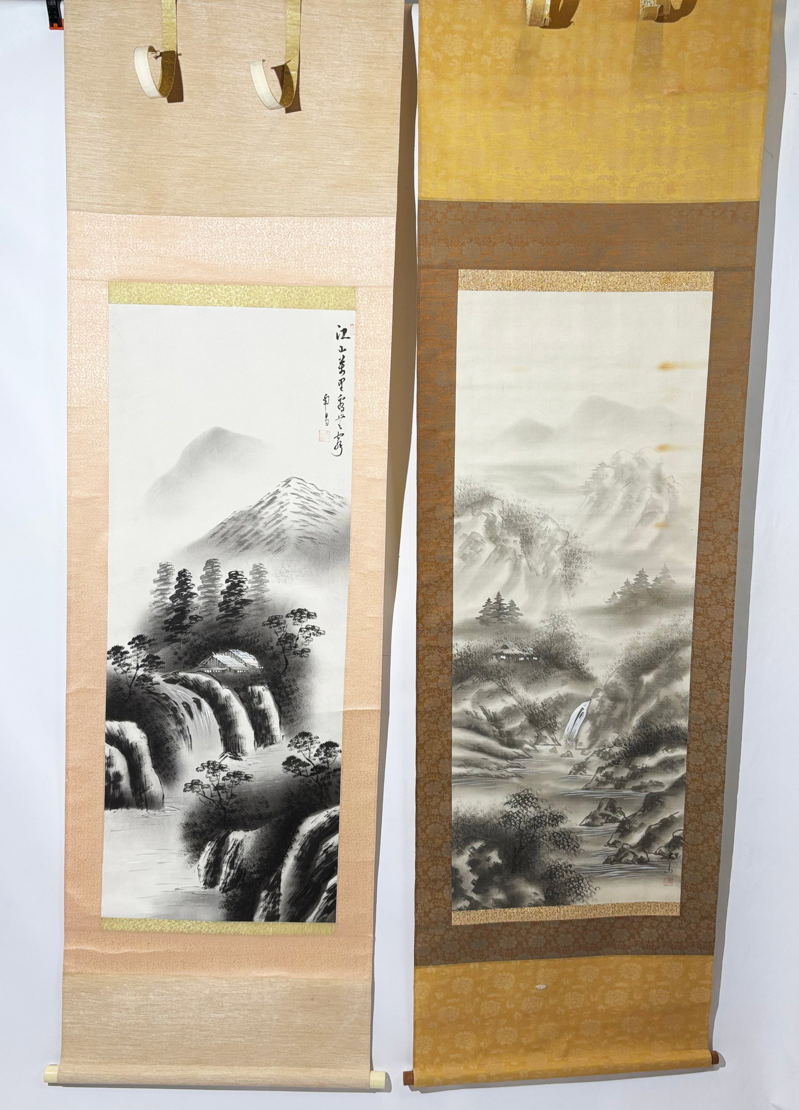 Two Japanese Ink Landscape Hanging Scrolls: Two vertical ink-on-paper hanging scrolls. Each depicts a misty mountain landscape featuring cascading waterfalls, woodland dwellings and flowing rivers. The right-hand scroll bears Japanese calligrap