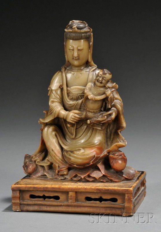 Soapstone Carving, China, late 19th century, finel: Soapstone Carving, China, late 19th century, finely carved as seated figure of Guanyin holding a boy, on a rectangular stand carved as a lotus pond, the stone of yellow green color with orange-red acc